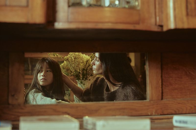 Two women, one Asian girl, are captured in a warm, intimate scene, reflecting in a mirror with a vase of white flowers.