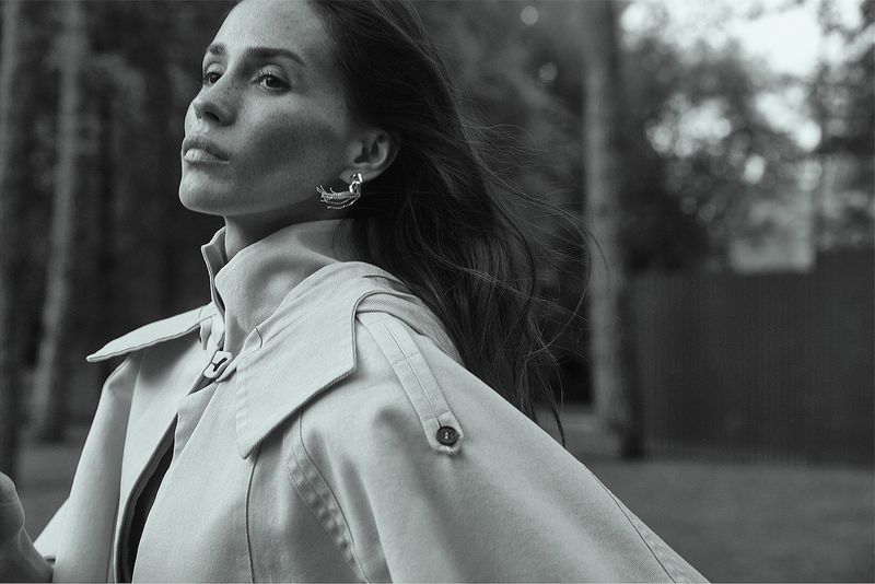Black and white portrait of an adult female wearing a trench coat and earrings in an outdoor park-like setting with trees.