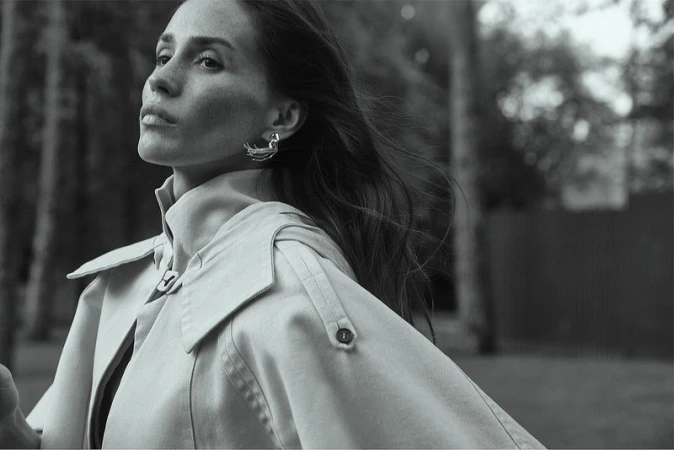 Black and white portrait of an adult female wearing a trench coat and earrings in an outdoor park-like setting with trees.