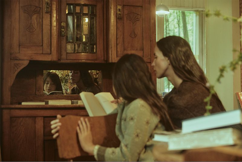 Two females of different generations are reading a book indoors with an antique cabinet in background and sunlight coming through a window.