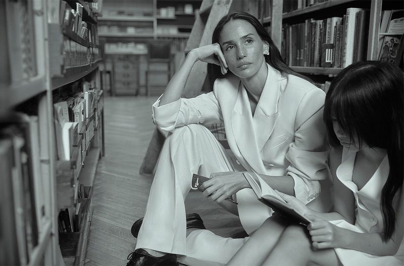 Two people, an adult and a child, sitting on the floor of a library surrounded by bookshelves in a black-and-white photograph.