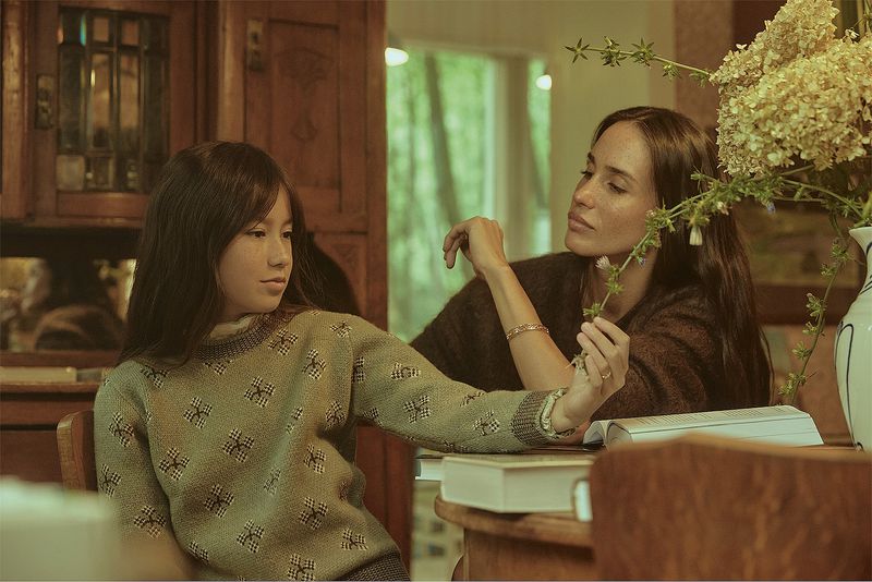 Portrait of an adult woman and a child indoors, seated at a wooden table with books and a vase of flowers in soft, natural light.