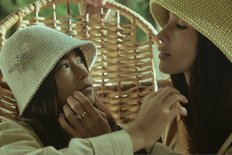 Two young women in crochet hats are captured in a close-up shot, with one woman gently touching the other's face in a natural outdoor setting.