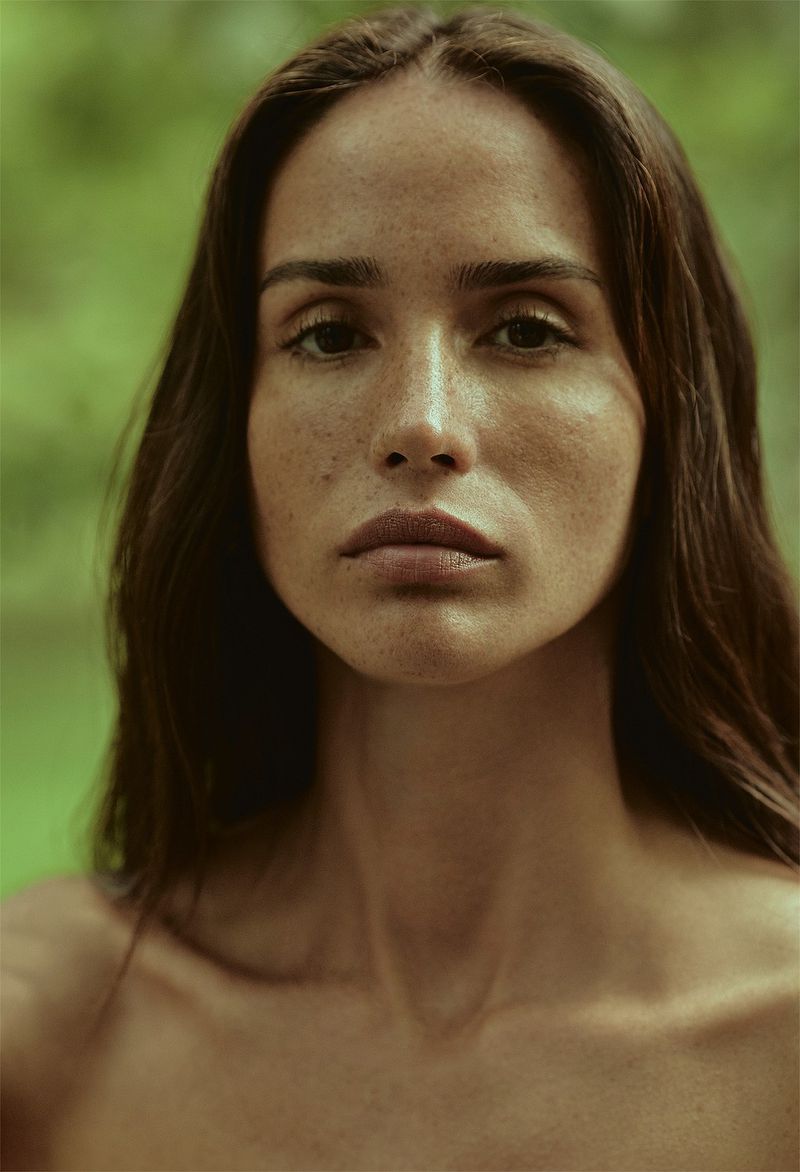 Portrait of a woman with dark hair and freckles, looking directly at the camera against a soft green, out-of-focus background. 