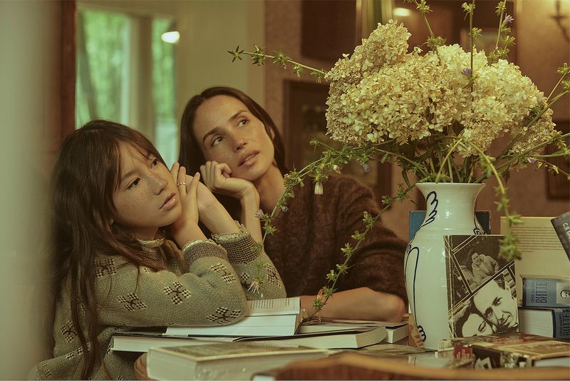 Mother and daughter pose at a table laden with books and a vase of white flowers in an indoor setting with soft, diffused lighting.