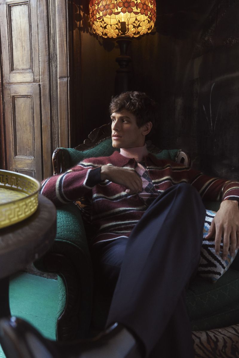 A male fashion model with dark hair is sitting in a chair indoors, wearing a striped sweater and trousers, lit by lamp light.
