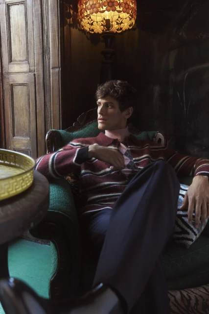 A male fashion model with dark hair is sitting in a chair indoors, wearing a striped sweater and trousers, lit by lamp light.