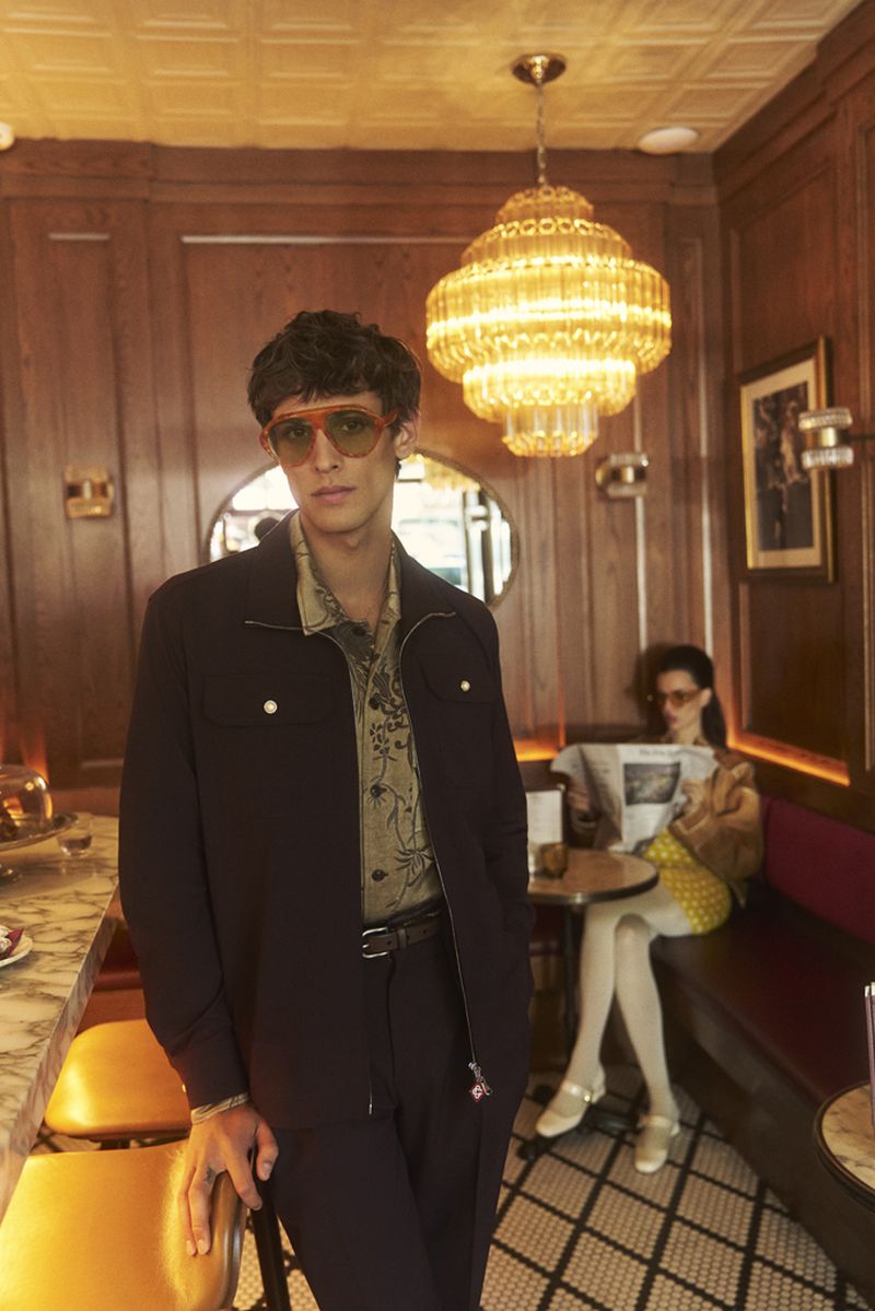 A stylish man in retro sunglasses and a jacket poses in a warm-toned diner setting, while a woman reads the newspaper in the background.