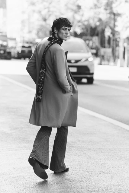 Black and white portrait of an adult male in a long coat and striped pants, walking on a city street with a car in the background.