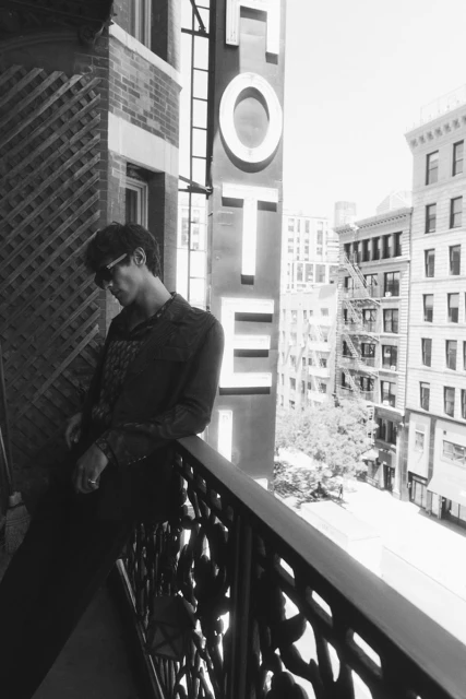 Monochrome portrait of a man on a balcony in front of a hotel sign and buildings in a city. He wears sunglasses and a patterned shirt.