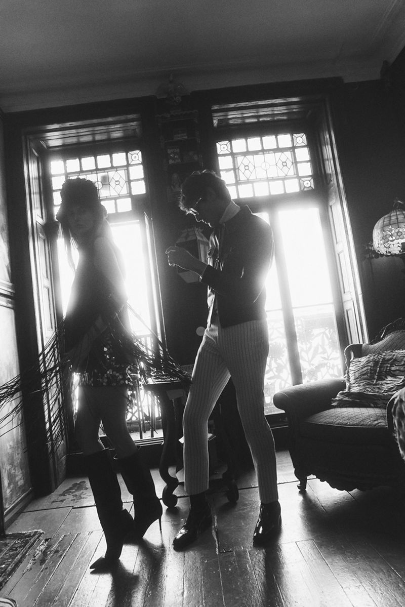 Monochrome shot of a fashionable couple standing indoors by a bright window, with details of their outfits visible.