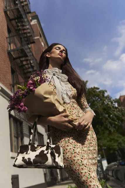 A woman in a floral dress, shot from a low angle, carrying a bouquet and handbag against an urban backdrop with fire escape.