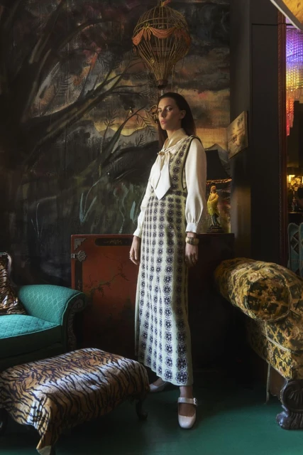A woman in a long, patterned dress and cream-colored blouse stands indoors, surrounded by vintage furniture and decor in a posed shot.