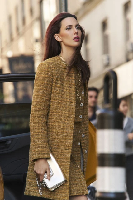 A woman with auburn hair walks on a city street wearing a tweed skirt suit, a gold chain, and carrying a metallic clutch in daytime.