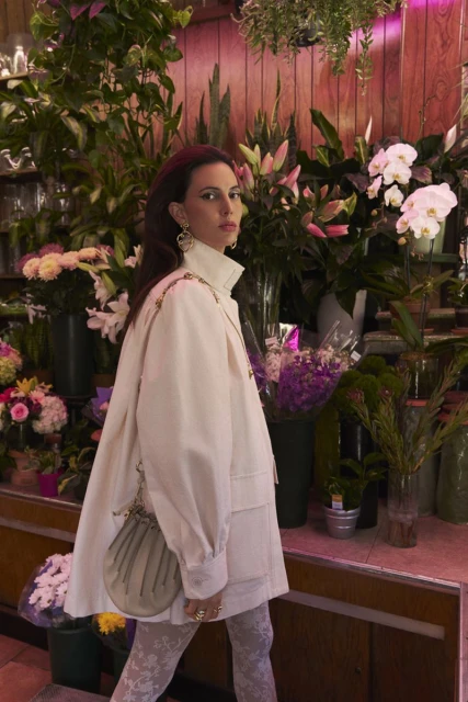 Adult female in a light jacket poses among orchids, lilies, and assorted plants in a flower shop with a pink wood-panel wall.