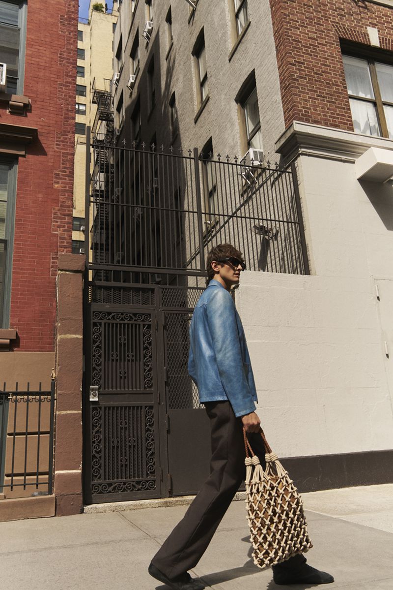 Adult male strides on city sidewalk, wearing a blue jacket, dark pants, and carrying a woven tote bag, captured in natural light.
