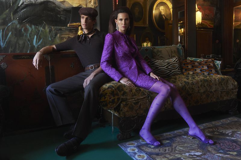 A fashionable couple poses indoors: a man in a cap and polo shirt sits next to a woman in a purple suit and patterned tights on a couch.