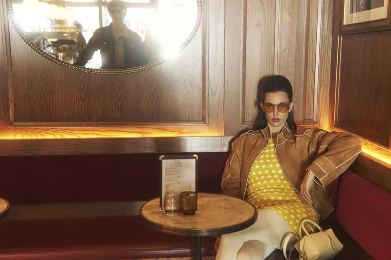 A woman in a yellow polka dot dress and brown jacket sits at a table in an indoor setting. A mirror reflects a person in the background.