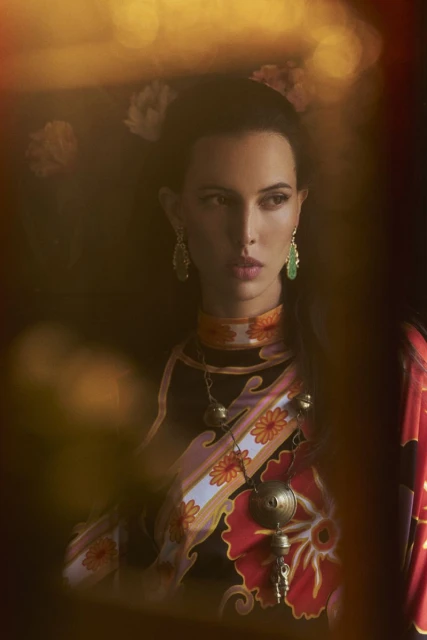 Portrait of an adult woman wearing floral and geometric print clothing and jewelry in a diffused, warm-toned indoor setting.