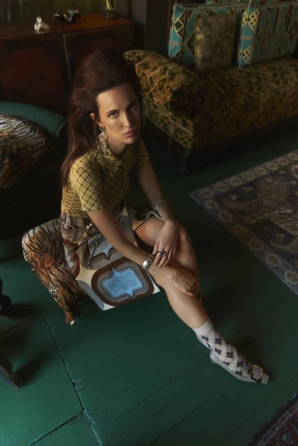 A woman with brown hair sits in an indoor setting, wearing a yellow and brown shirt and patterned skirt. Cushions and a rug are visible.