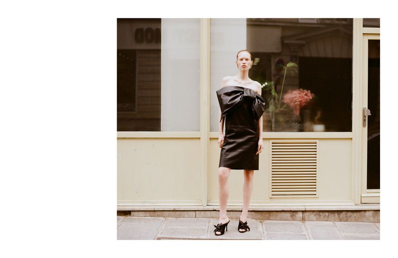A slender fashion model poses outside in a black satin dress adorned with a large bow, accessorized with black heeled mules and minimal jewelry.