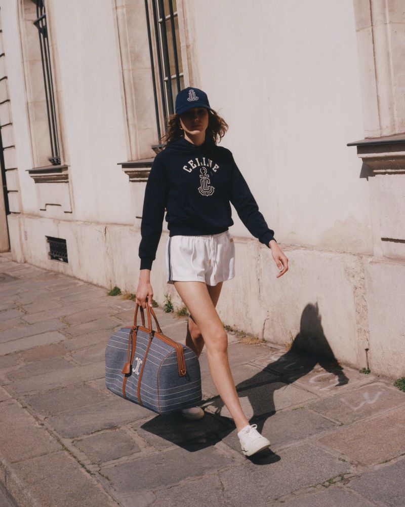 A woman walks on a city street, wearing a navy hoodie and cap with logo, white shorts, white sneakers, and carrying a duffel bag.