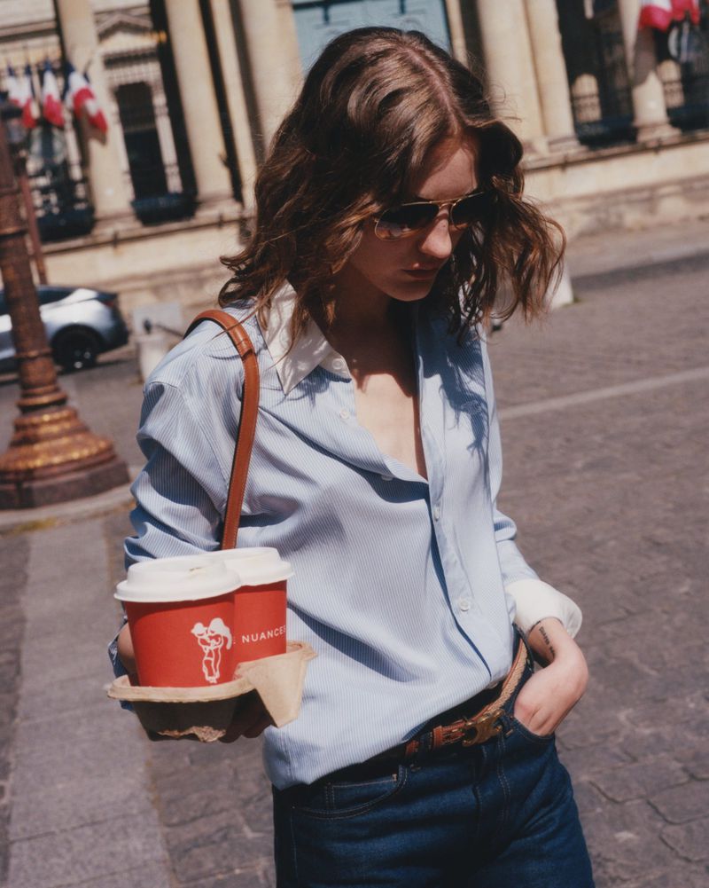 Adult female in jeans and shirt holding coffee on urban street with building and French flags in the background during daytime.