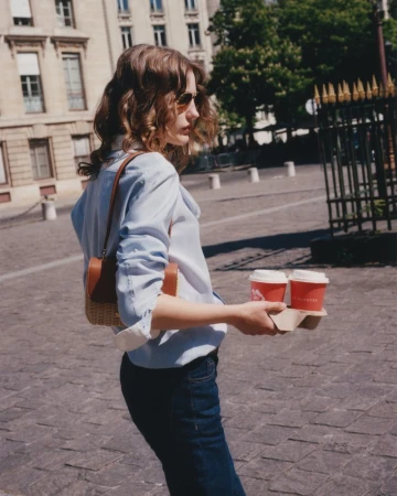 Woman in light-blue shirt carrying two coffee cups on a paved street in a European city on a sunny day. Buildings and trees in background.