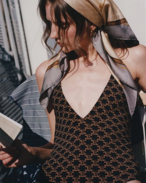 Woman in patterned one-piece swimsuit and headscarf reading a book, sitting outdoors in bright sunlight. Fashion, style, summer.