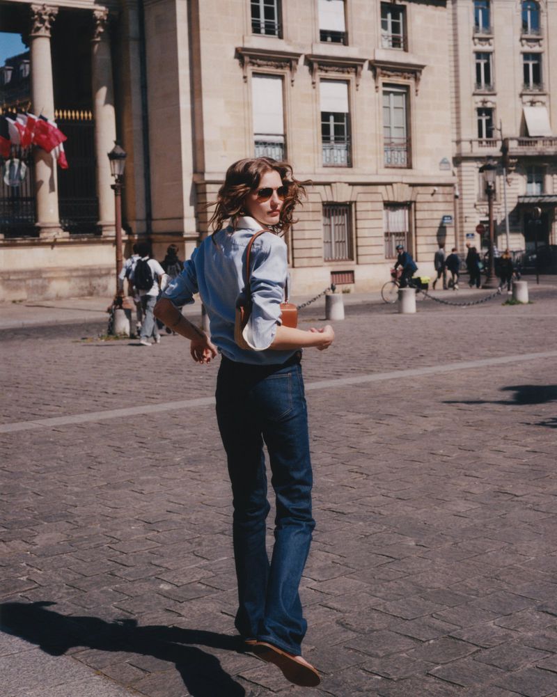Woman walking on cobblestone street with architecture in the background. Wearing a button-down shirt, jeans, and sunglasses.