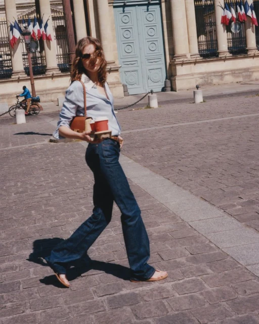 Stylish woman in blue jeans walks across a plaza in front of a European building, carrying a shoulder bag and coffee.