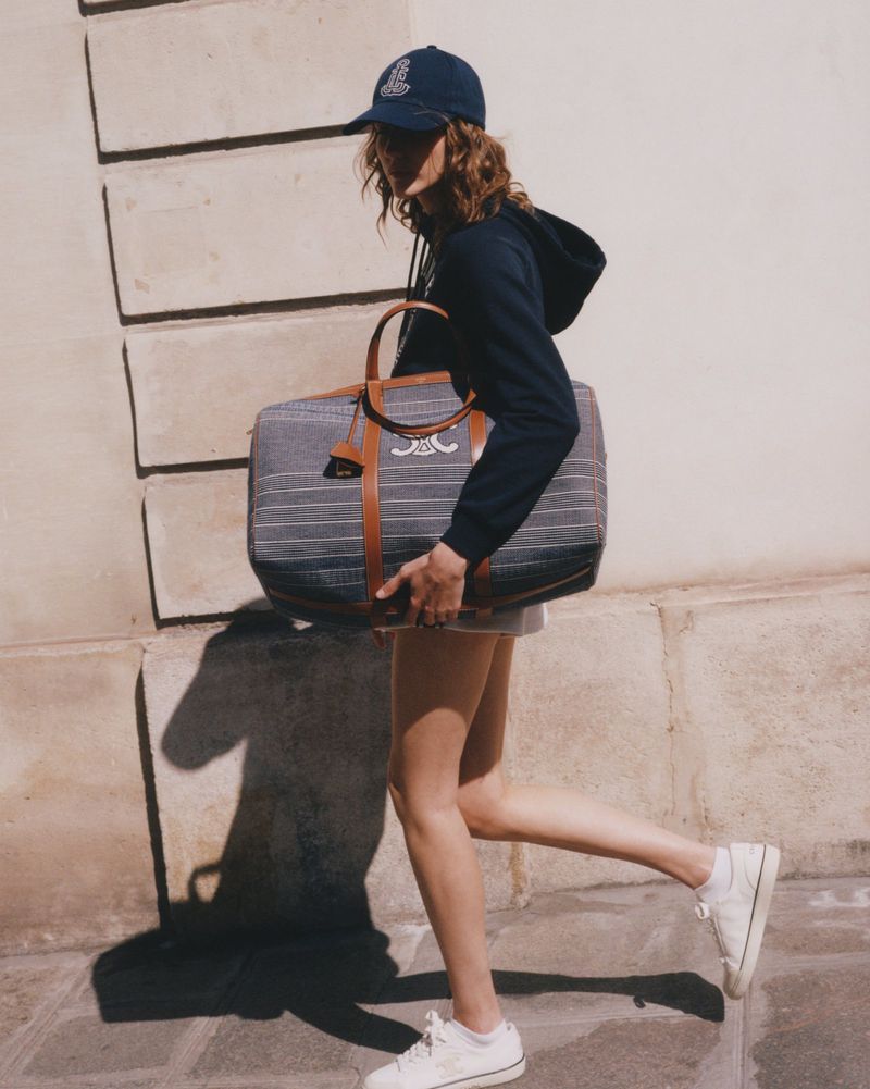 Fashionable woman in a baseball cap, hoodie, and sneakers carries a striped duffel bag walking on a sunny day in an urban setting.