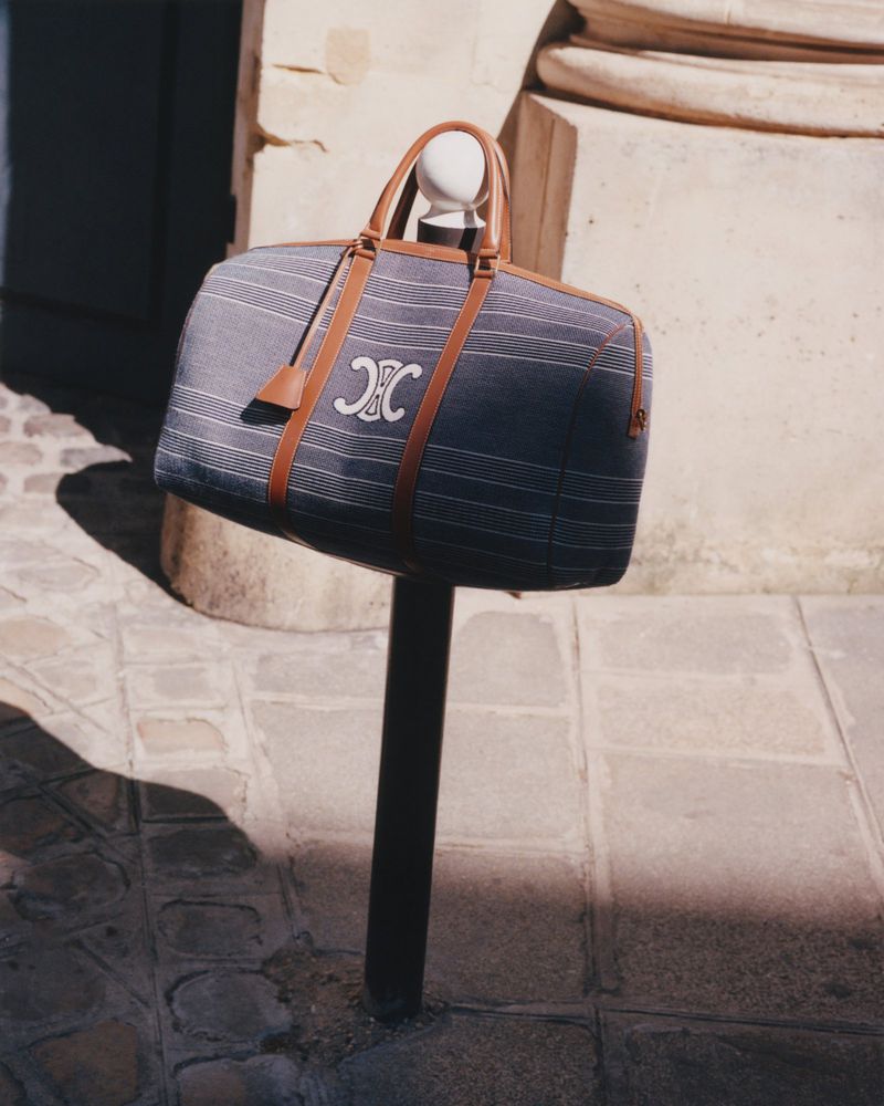 Striped fabric duffel bag with leather trim featuring a white 'Triomphe' logo, placed on a metal post in an outdoor setting.