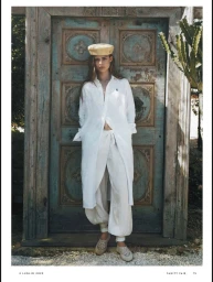 Model wearing a white shirt dress, loose trousers, and beret standing before an ornate turquoise wooden door.