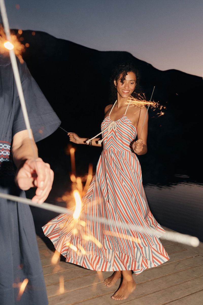 Woman in red and white striped dress dancing with sparklers on a wooden deck at dusk with mountains in the background and two people.