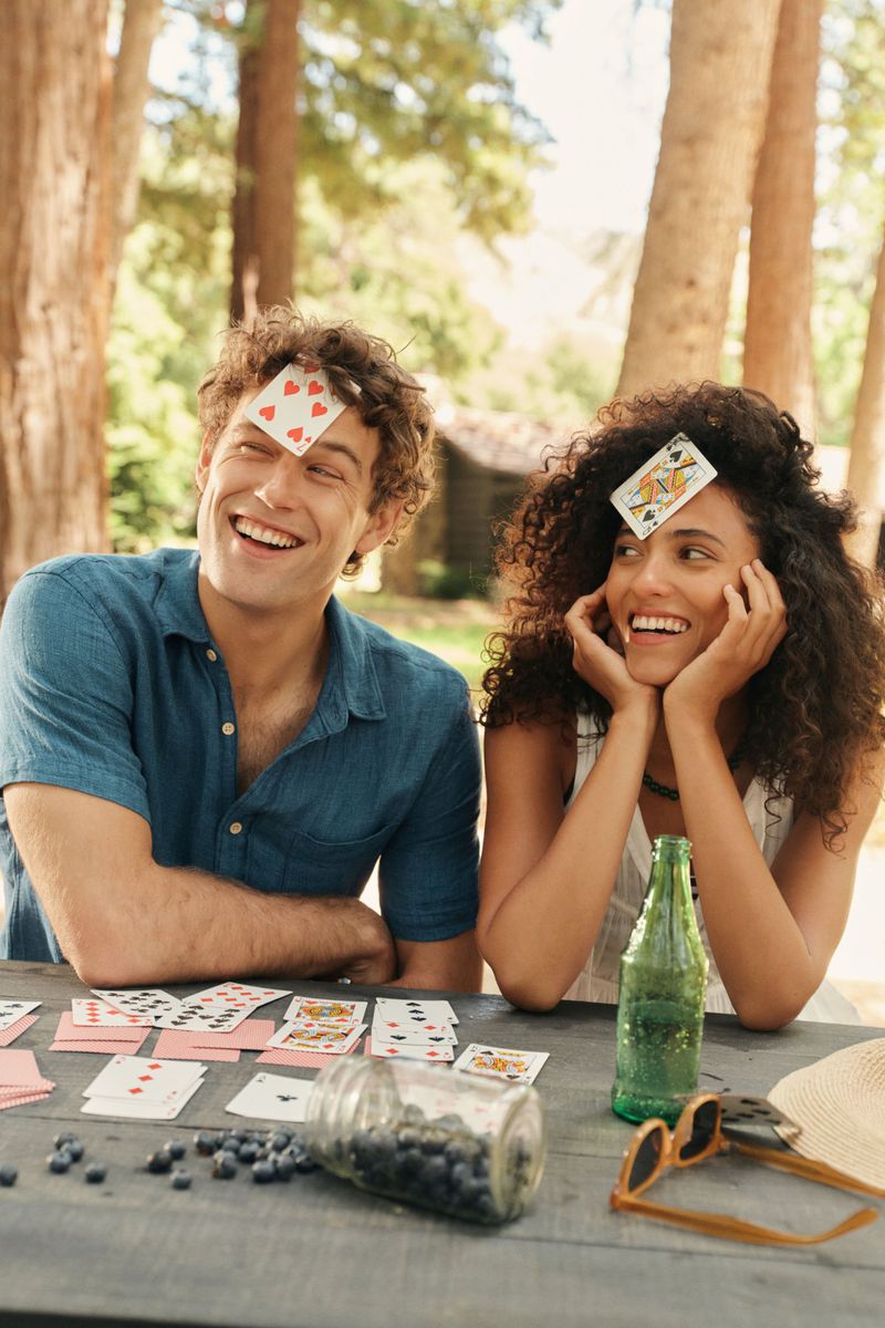 A happy couple sits outdoors with playing cards on their foreheads, sharing a lighthearted moment amidst a playful card game in natural light.