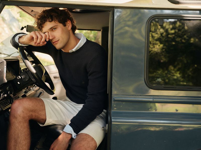 Handsome adult male poses in a vintage car on a sunny day, wearing a navy sweater and beige shorts, set against a blurred outdoor background.