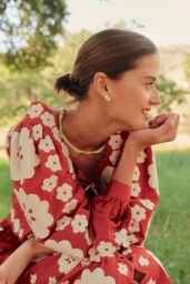 Smiling woman in a red and cream floral long-sleeve dress and gold necklace outdoors in a sunlit grassy field.