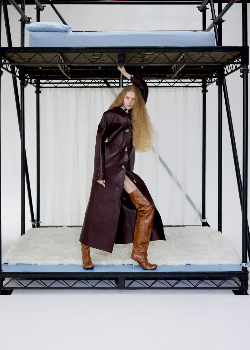Fashion shot of a long-haired woman wearing a brown coat and patterned boots, standing on a metal bed frame with a white background.