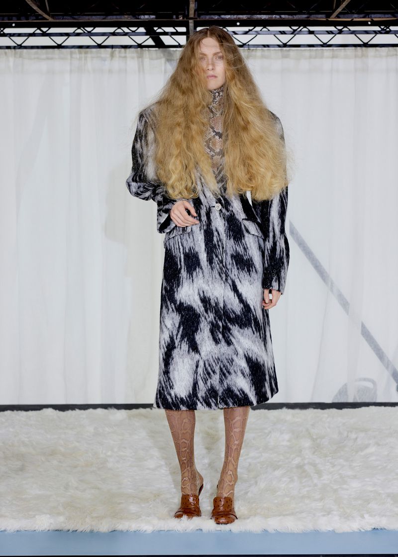 Model with voluminous crimped hair wearing a black and white patterned coat standing on a white shag rug in front of a curtain.