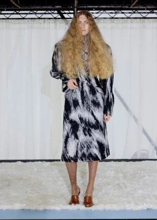 Model with voluminous crimped hair wearing a black and white patterned coat standing on a white shag rug in front of a curtain.