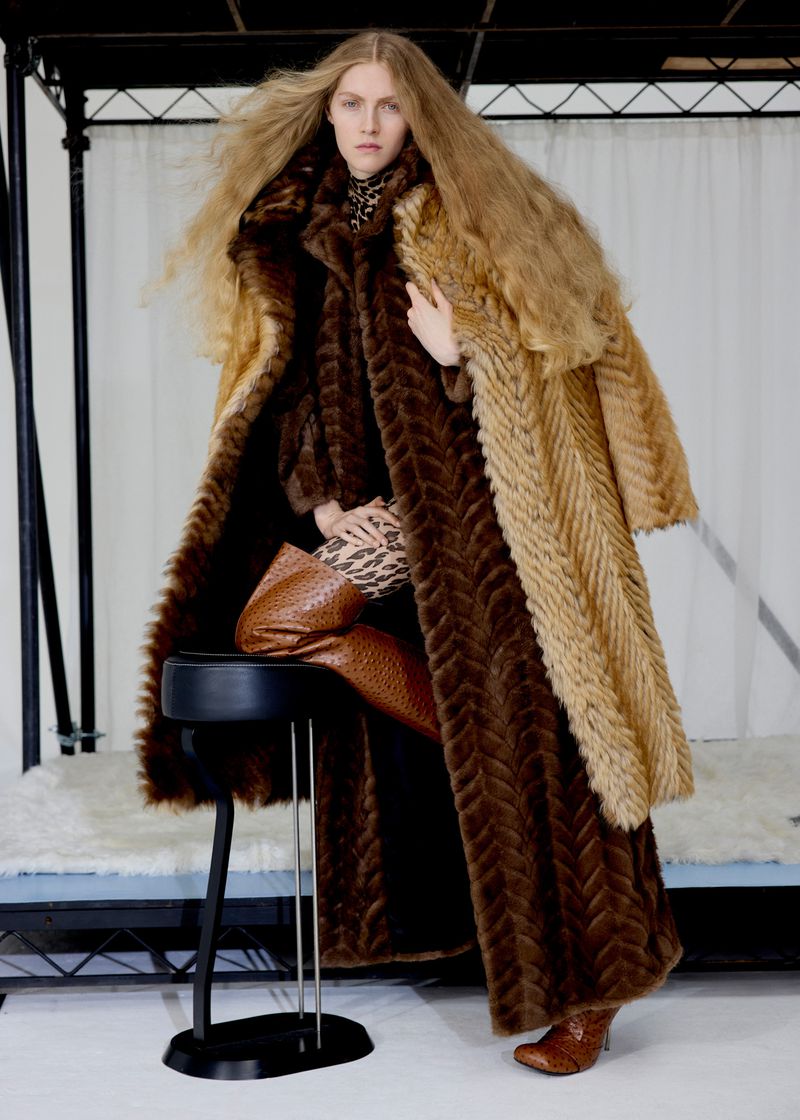 A fair-skinned woman with long blond hair poses, wearing a long patterned brown and blonde fur coat and reptile-print boots.