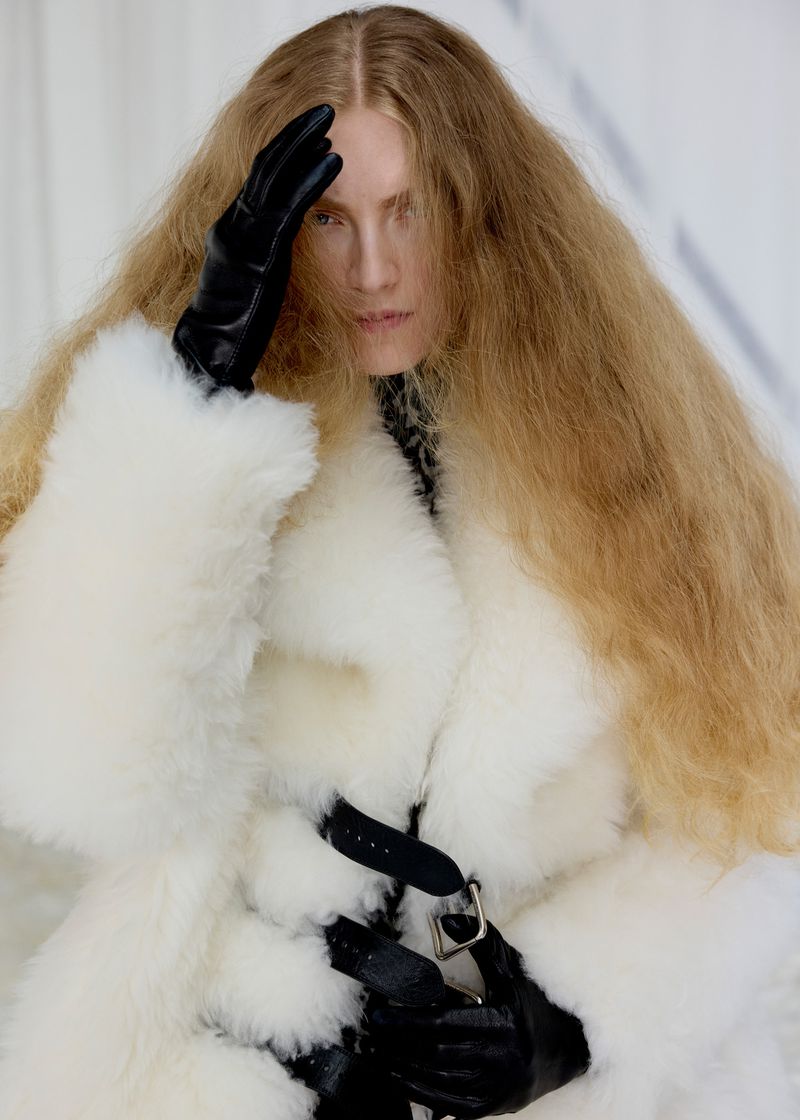 A blonde model with long wavy hair wears a white fluffy coat, black gloves, and a harness-style belt in a minimalist indoor setting.
