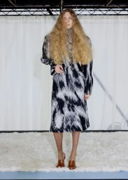 Model with voluminous crimped hair wearing a black and white patterned coat standing on a white shag rug in front of a curtain.