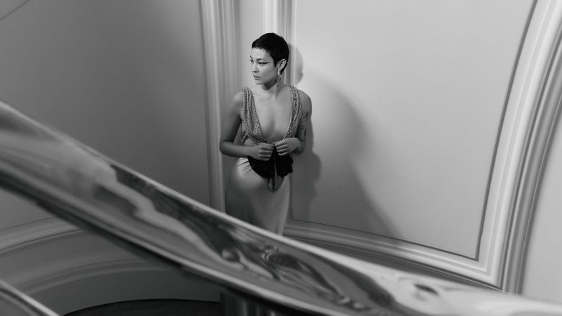A woman with a pixie cut poses in a low-cut dress on a staircase, creating a high-fashion black and white portrait, indoors.
