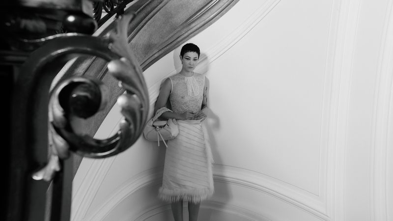 Monochrome fashion shot of woman in lace top and skirt with handbag posing near ornate staircase railing in an indoor environment.