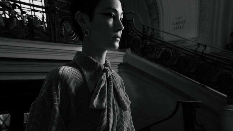 Monochrome portrait of a woman in patterned top standing on stairs with ornate railing. Focus on the subject, with soft lighting.