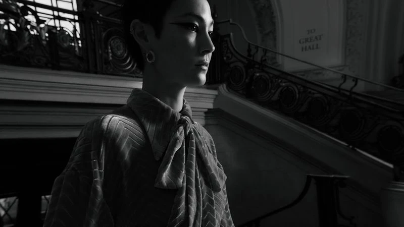 Monochrome portrait of a woman in patterned top standing on stairs with ornate railing. Focus on the subject, with soft lighting.