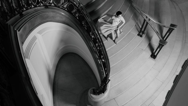 Monochrome image of a stylish adult female descends a spiral staircase, wearing a flowing dress with a hand on the railing.