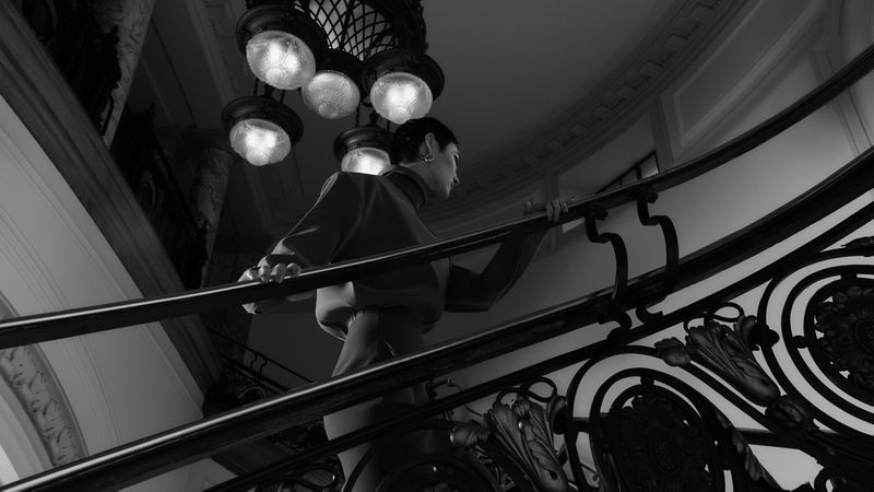 A woman climbs an ornate staircase with black metal railings inside a building with a high ceiling and decorative lighting fixtures.